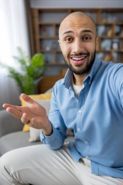 Genç kel bir adam, modern oturma odasındaki kanepesinden gelen video görüntülerinde gülümsüyor ve el kol hareketi yapıyor. Günlük mavi gömlekli, kendinden emin ve kameraya çekici.