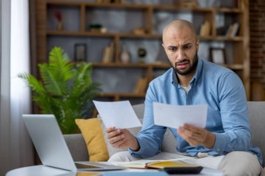 Evdeki bir kanepede oturan genç adam, mali belgeleri, faturaları ve evrak işlerini elinde tutarken endişeli ve sıkıntılı görünüyor.