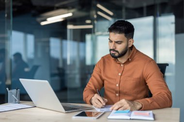 Çağdaş bir ofis ortamında üretken bir iş gününde masasında dizüstü bilgisayar ve tablet bulundurarak not almaya odaklanan genç işadamı.