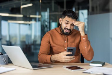 Stresli genç işadamı cep telefonunda kötü bir mesaj okurken hayal kırıklığına uğramış ve üzgün hissediyor, modern ofis ortamında bir sorun ya da hata yaşıyor