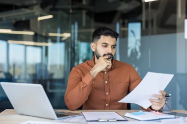 Ofis masasında oturan odaklanmış işadamı, düşünceli bir şekilde belgeleri analiz ediyor ve bir dizüstü bilgisayar üzerinde çalışırken verileri inceliyor, yeni bir iş stratejisine konsantre oluyor.