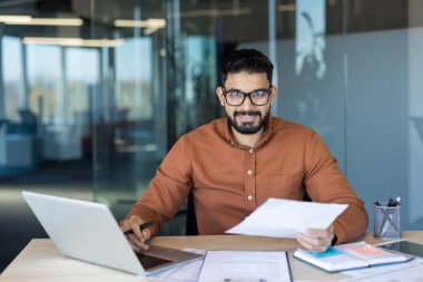 Hintli işadamı bir ofis masasında oturuyor, elinde belgelerle kameraya gülümsüyor ve bir dizüstü bilgisayarla çalışıyor, modern bir çalışma alanında görevleri ve evrak işlerini yönetiyor.