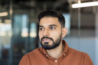 Sakallı ve ciddi ifadeli genç adam, derin düşünceler içinde, modern ofis ortamında planlama, görme, düşünme ve geleceğe odaklanma kavramlarını temsil ediyor.