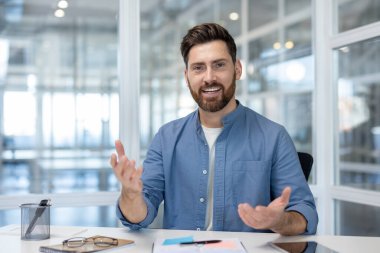 Çalışma masasından aktif olarak bir video görüşmesine veya çevrimiçi konferansa katılan genç işadamı konuşurken, konuşurken, iletişim ve bağlantı kurarken, yaygın bir şekilde gülümsüyor ve işaret ediyor