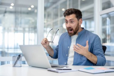 İşadamı, elinde gözlüklerle dizüstü bilgisayarının ekranına bakıyor. Beklenmedik bilgilere aşırı derecede şaşırmış ve güvensiz bir şekilde bir ofis masasında oturuyor.