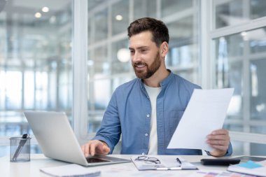 Ofisteki genç sakallı girişimci, dizüstü bilgisayarla çalışırken gülümsüyor ve ticari belgeleri inceliyor, üretkenliği, güveni ve modern profesyonel başarıyı aktarıyor.