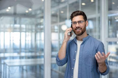 Genç işadamı mutlu bir ifade ve jestle cep telefonuyla konuşuyor. Çağdaş bir şirket ortamında iş görüşmesi sırasında pozitif ve kendinden emin hissediyor.