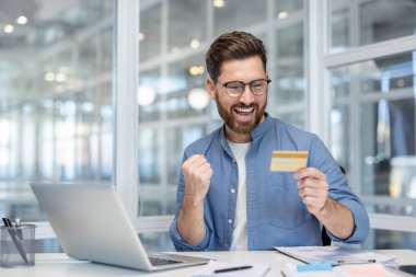 Gözlüklü sakallı bir adam elinde bir kredi kartı tutarken ve bir dizüstü bilgisayara bakarken, ev ofisinden veya modern çalışma alanından online alım başarıyla tamamlıyor.