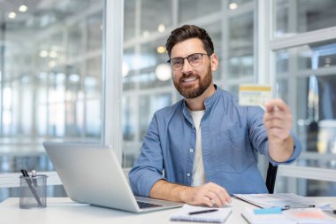 Bilgisayarı online ödeme için kullanırken elinde kredi kartı tutan gülümseyen adam, modern bir ofiste güvenli e-ticaret, dijital bankacılık ve uygun uzaktan işlemleri temsil ediyor.