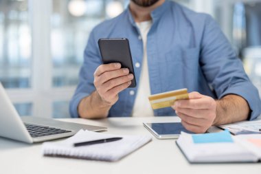 Elinde akıllı telefon ve kredi kartı olan bir adam, bir ofis masasından online ödeme ve e-ticaret işlemleriyle ilgileniyor, modern dijital finans ve alışveriş olanakları sunuyor.