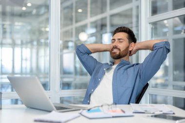 Ofis sandalyesinde gözleri kapalı, rahatça gülümseyen, rahatlamış, tatmin olmuş ve kendinden emin genç bir profesyonel.