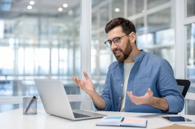 Çevrimiçi bir toplantı sırasında erkek profesyonel iletişimi, modern bir ofis ortamında dizüstü bilgisayar kullanarak video görüşmesinde el kol hareketi yapmak.