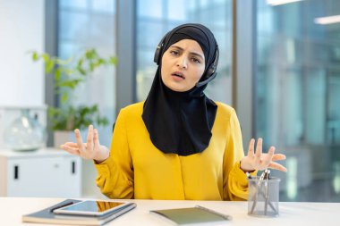 Ofis masasında kulaklık takan Müslüman bir kadın. Kafası karışmış ve hayal kırıklığına uğramış bir ifadeyle doğrudan kameraya bakıyor. Çağrı merkezinde çalışırken kafasını karıştırmak için ellerini açtı.