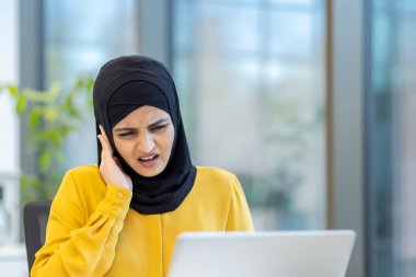 Başında laptopuna odaklanan ve kulak ağrısı ya da baş ağrısı çeken müslüman bir kadın ofis ortamında kederli bir ifadeyle kulağına dokunuyor.