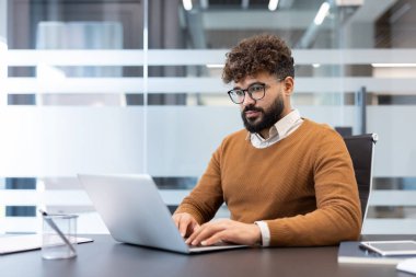 Gözlüklü ve sakallı genç bir işadamı modern, parlak bir ofiste, çağdaş bir masada özenle çalışıyor, görevlerine odaklanıyor ve üretkenliğini gösteriyor.