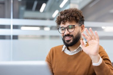 Kıvırcık saçlı ve gözlüklü gülümseyen adam uzak bir online video görüşmesi sırasında el sallıyor, modern ofis alanından meslektaşları ve arkadaşlarıyla bağlantı kuruyor.