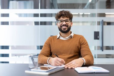 Modern bir ofis masasında oturan sakallı ve gözlüklü genç adam, bir not defterine not tutarken kameraya gülümsüyor, üretkenliği ve başarıyı simgeliyor.