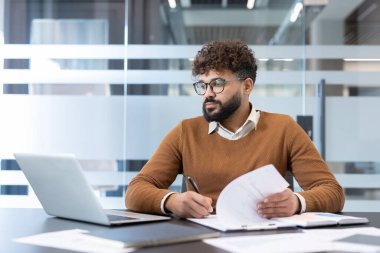 Young confident businessman with beard and glasses actively working at an office desk, reviewing documents, reading papers, and making notes while using a laptop
