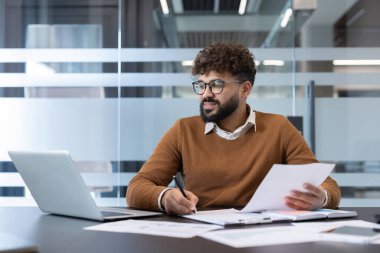 Modern bir ofis masasında çalışan genç bir işadamı, belgeler üzerinde çalışır, notlar alır ve bir dizüstü bilgisayarda çalışır, profesyonel bir ortamda araştırma, planlama ve üretkenliğe odaklanır.