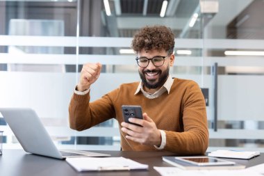 Gözlüklü hevesli genç adam başarıyı kutlarken yumruğunu sıkar ve modern bir ofiste akıllı telefonuna bakar heyecanlı ve kazandığını hisseder.