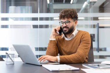 Genç iş adamı birden fazla iş yapıyor, dizüstü bilgisayarda yazarken telefon görüşmesi yapıyor, modern ofis ortamında cam duvarlı masasına gülümsüyor.