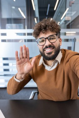 Kıvırcık saçlı ve sakallı gülümseyen adam kameraya bakıyor, elini sallıyor ve modern ofis çalışma alanından video konferans yoluyla iletişim kuruyor, uzaktan çalışma ve iş iletişimini temsil ediyor.