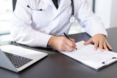 Masasında steteskoplu beyaz önlüklü bir doktor klinik konsültasyon sırasında laptopun yanındaki panoya reçete ve hasta notları yazıyor.