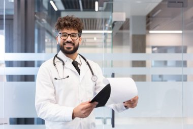 Laboratuvar önlüğü ve steteskop giyen genç bir erkek doktor modern bir klinik veya hastane ortamında durup kameraya bakıp gülümserken elinde tıbbi belgeler tutuyor.