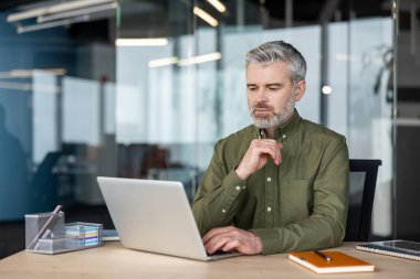 Olgun yönetici modern bir ofiste bir masada oturuyor, dizüstü bilgisayara odaklanıyor, yeni iş stratejileri planlıyor ve karmaşık problemleri düşünceli bir ifadeyle çözüyordu.