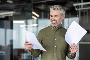 Modern bir ofiste gri saçlı deneyimli bir işadamı, raporları okuyup analiz ederken gülümsüyor, güven, başarı ve profesyonel liderliği aktarıyor.