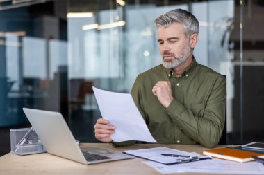 Olgun yönetici evrak işleri ve veri analizine odaklandı, bir dizüstü bilgisayar ve çeşitli belgelerle modern bir ofis masasında oturmak, gayretin ve kurumsal sorumluluğun somutlaştırılması