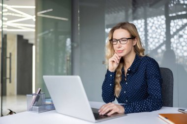 Gözlüklü genç profesyonel bir kadın, gümüş bir dizüstü bilgisayarla modern bir ofis ortamında çalışırken, kendine güvenen ve üretken bir şekilde masa başında daktilo kullanıyor.