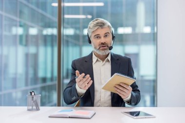 Deneyimli iş adamı çevrimiçi müşteri desteği ya da danışmanlık hizmeti sunuyor, kulaklık takıyor ve not defteri tutuyor, modern bir ofis ortamında video görüşmesi yapıyor.