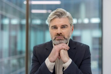 Gri sakallı, şık bir saç kesimi olan takım elbiseli, düşünceli ve odaklanmış bir ifadeyle kameraya bakan olgun bir adam.