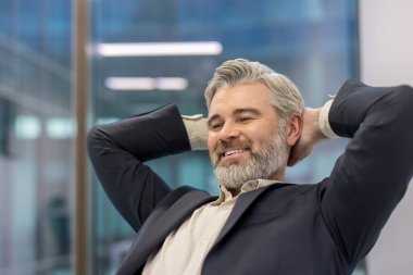 Olgun bir yönetici gülümseyerek ellerini başının arkasına koyup arkasını yaslar ve modern ofis ortamında başarıyı kutlar.