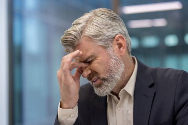 Takım elbiseli, gri sakallı, baş ağrısı ve göz yorgunluğundan gözleri kapalı, ofis ortamında stres, yorgunluk ve yorgunluk taşıyan olgun bir işadamı.