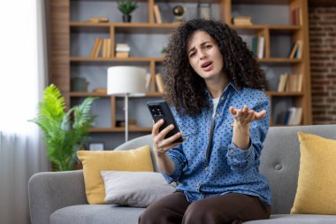 Woman with curly hair sitting on sofa, looking perplexed and making a gesturing hand, clearly expressing frustration and confusion while holding and looking at her smartphone