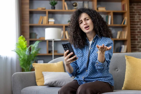 Woman with curly hair sitting on sofa, looking perplexed and making a gesturing hand, clearly expressing frustration and confusion while holding and looking at her smartphone