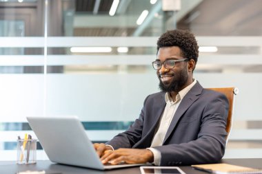 Gözlüklü siyahi işadamı dizüstü bilgisayarında gülümseyip daktilo kullanıyor. Çağdaş, profesyonel bir ofis ortamında masa başı işleriyle meşgul.