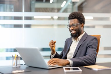 İş adamı iyi haberler alıyor ya da başarılı oluyor. Çağdaş ofis ortamında dizüstü bilgisayara bakarken yumruğunu sallıyor ve gülümsüyor.