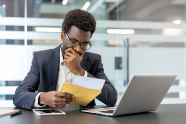 Stresli bir mektup ya da belge okuduktan sonra endişeli görünen bir işadamı, eli çenesinde sarı bir zarf tutuyor, bir bilgisayar ve akıllı telefonuyla bir ofis masasında oturuyor.