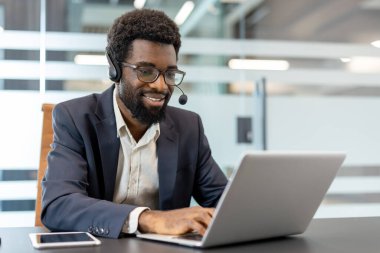 Mikrofon ve gözlüklü bir kulaklık takan Afrikalı bir adam, bir masada oturuyor, dizüstü bilgisayarla yazarken gülümsüyor, modern ofis ortamında müşteri hizmetleri sunuyor.