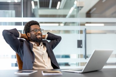 Afrikalı işadamı ellerini başının arkasına koyup masasında rahat bir şekilde oturuyor. Dizüstü bilgisayarındaki işi bitirdikten sonra gülümsüyor ve tatmin oluyor.