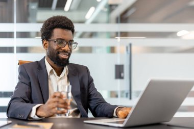 Akıllı takım elbiseli bir iş adamı, modern bir şirket masasında dizüstü bilgisayarla çalışırken, elinde bir bardak su, kendinden emin ve uzaktan çalışma sırasında odaklanmış bir şekilde gülümsüyor.