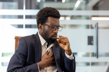 Afrikalı Amerikalı iş adamı hasta hissediyor, öksürürken ağzını kapatıyor, bir eliyle göğsüne bastırıyor ve modern bir ofiste acı ya da rahatsızlık hissediyor.
