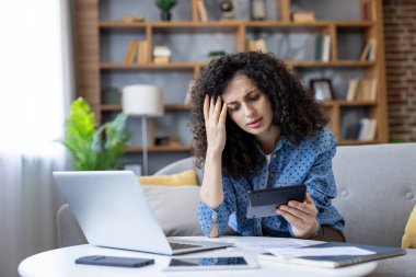 Kadın, evde hesap makinesi ve dizüstü bilgisayarla ev giderlerini, faturaları ve belgeleri analiz ederken stresli ve endişeli hissediyor ve mali zorluklar yaşıyor.