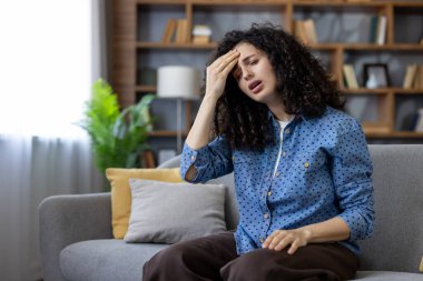 Şiddetli baş ağrısı ya da stresten muzdarip genç bir kadın, modern bir oturma odasında rahat gri bir koltukta oturuyor, gözleri kapalı ve rahatsız edici bir şekilde şakaklarını tutuyor.