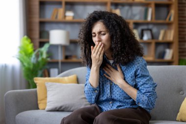 Genç bir kadın kendini iyi hissetmiyor ve grip ya da soğuk algınlığı belirtileri gösteriyor. Evdeki bir kanepede otururken eline öksürüyor. Rahatsızlık ve hastalık belirtileri gösteriyor.