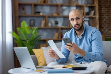 Evdeki bir kanepede oturan serbest çalışan erkek, uzun bir banknot tutarken stresli görünüyor ve akıllı telefonunu mobil bankacılık veya bütçe için kullanıyor, yakınlardaki bir dizüstü bilgisayarla kişisel finansman yönetiyor.