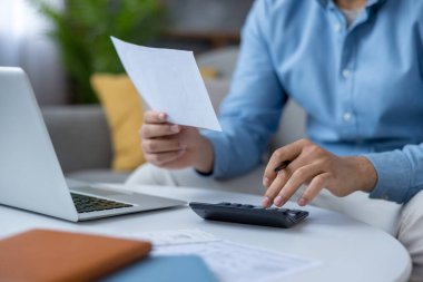 Elinde kağıt bir belge tutan ve dizüstü bilgisayarlı bir masada otururken hesap makinesi kullanan, kişisel finansman yöneten, aylık giderleri karşılayan ve evdeki faturaları gözden geçiren bir adam.
